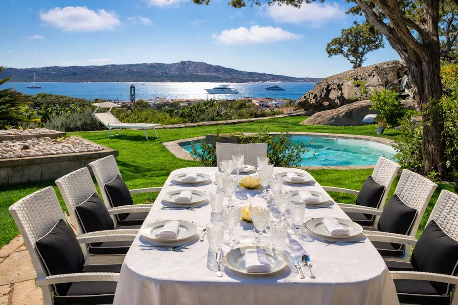 Outdoor dining table set with white linens, plates, and glasses, surrounded by white chairs with black cushions. Pool, ocean, and mountain views in the background.