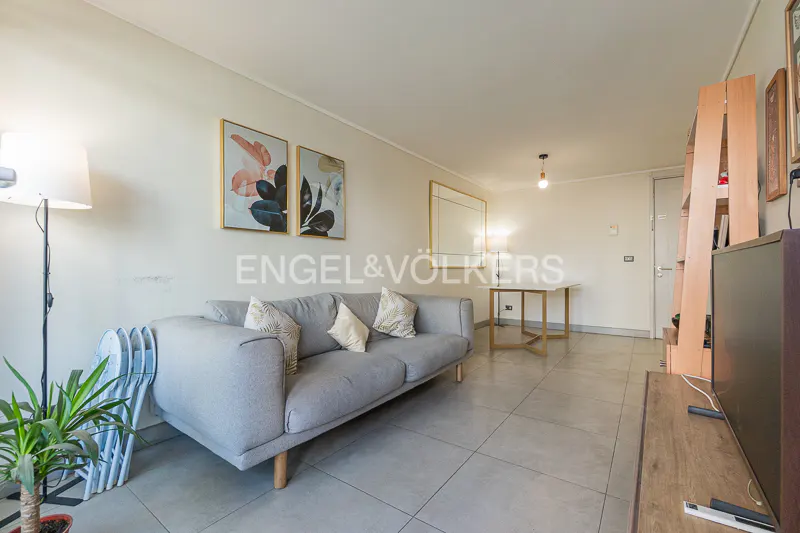 Living room with a gray sofa, patterned pillows, and framed art on the wall. A floor lamp and plant are near the sofa. A table is in the background.