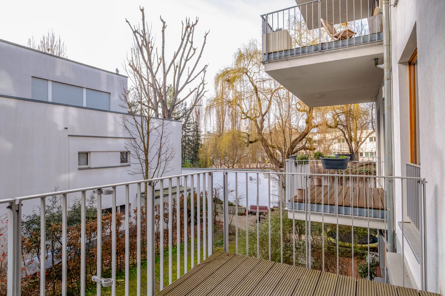 View from a wood-decked balcony with white railings, overlooking a pond and trees. A white building is visible to the left.