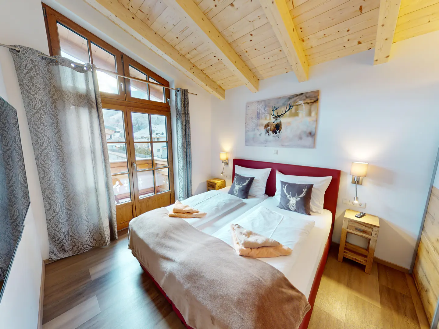 Bedroom with a red bed frame, white linens, deer pillows, and a wooden ceiling. A painting of deer hangs above the bed. Curtains frame a window with a view.