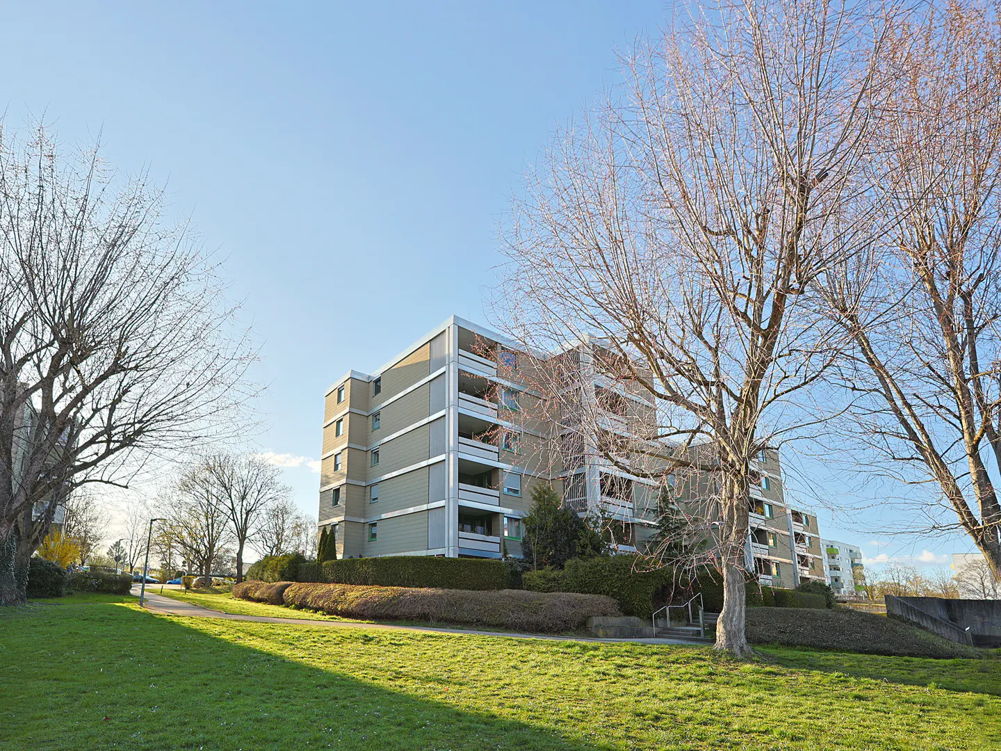 Exterior view of a multi-story apartment building with balconies, green lawn, and bare trees under a blue sky.