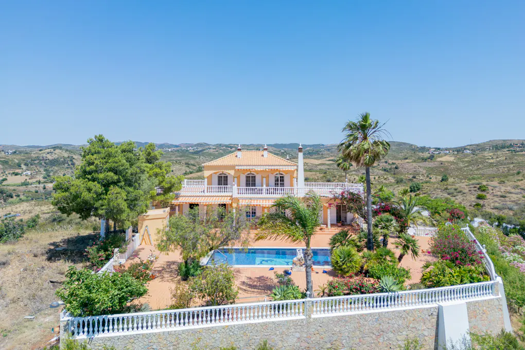 Aerial view of a yellow two-story villa with a swimming pool, palm trees, and white railings, set in a hilly landscape.