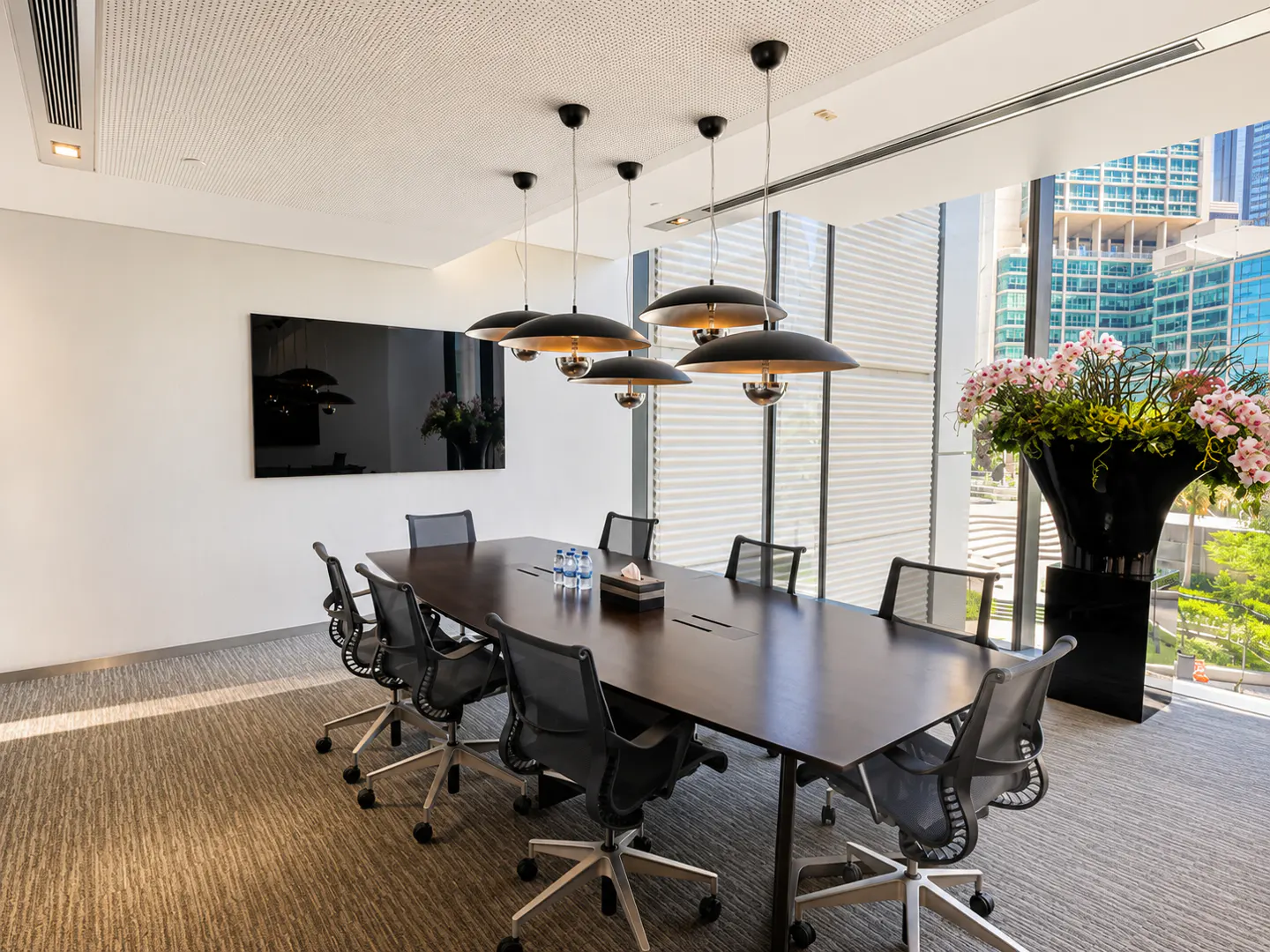 Modern boardroom with a long dark table, black mesh chairs, pendant lights, and a large planter with pink orchids.