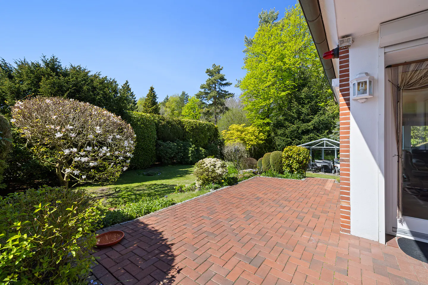 A brick patio leads to a lush green garden with trees, bushes, and a gazebo under a clear blue sky.