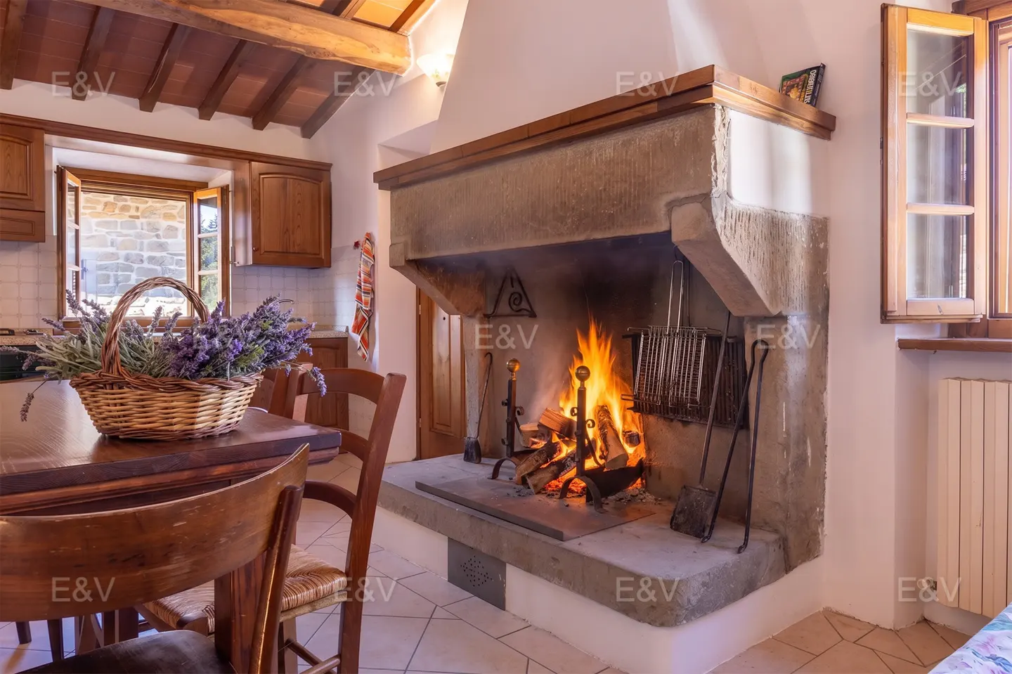 A rustic kitchen with a stone fireplace ablaze, wooden table with lavender basket, and open window.