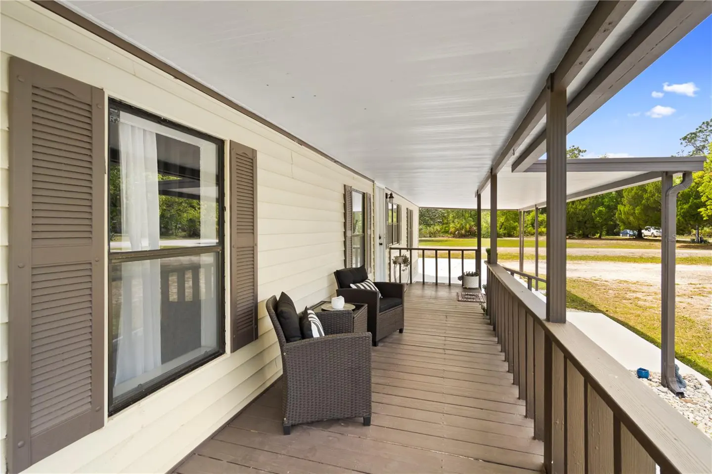 Covered porch with two dark wicker chairs and a small table. The house is light yellow with brown shutters and trim. A railing surrounds the porch.