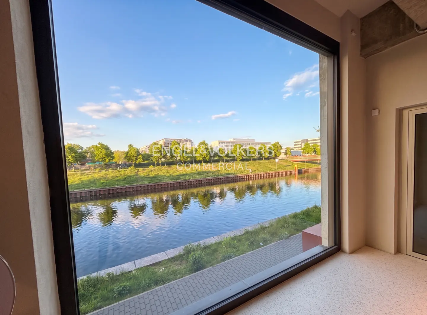 View from a commercial space window overlooking a river, green trees, and buildings under a blue sky.