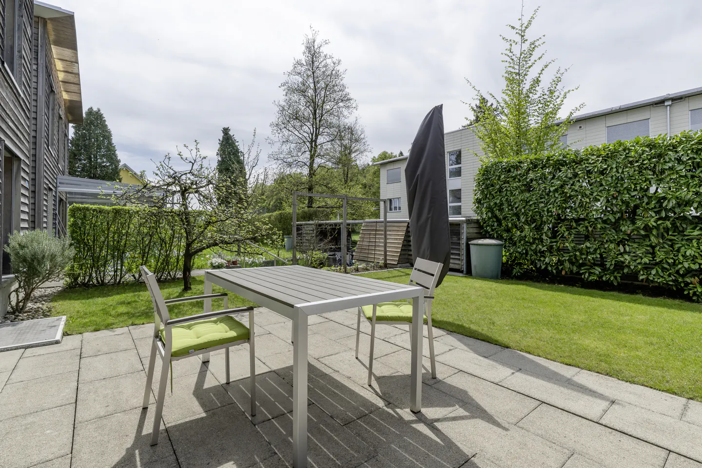 Patio with a gray table and two chairs with green cushions on a stone patio, with a green lawn and trees in the background.
