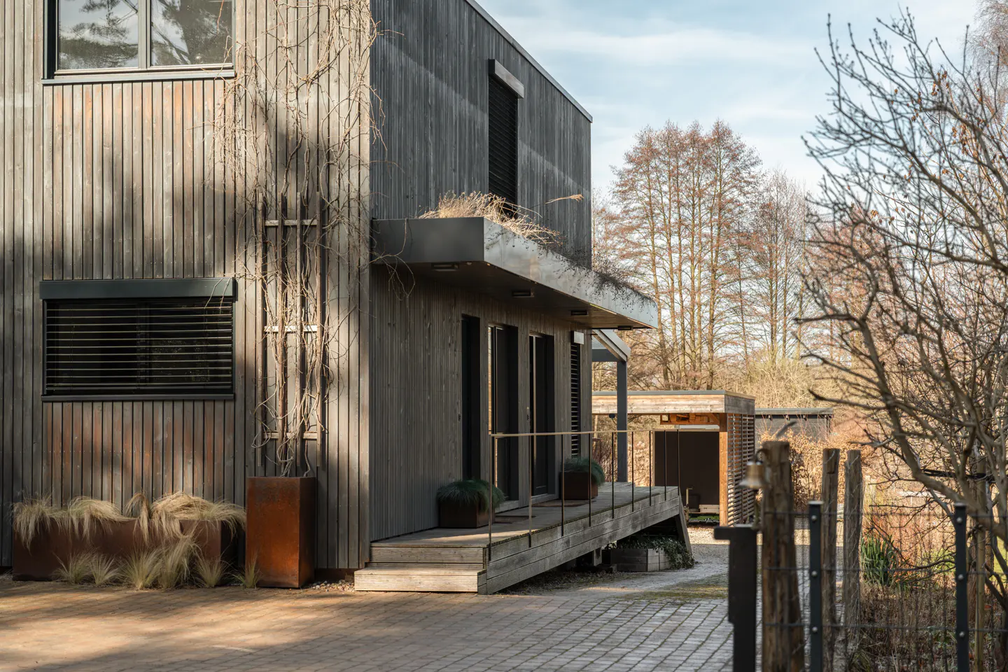 Modern home with gray wood siding, a wooden porch, and a brick driveway. Trees are visible in the background.