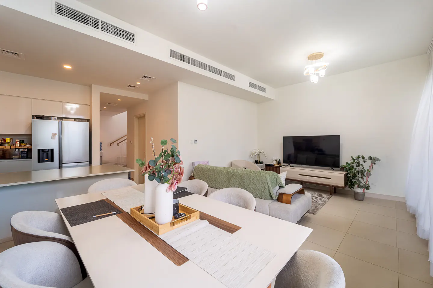Open-concept living space with a white dining table, chairs, and a vase of flowers. A sofa, TV, and kitchen are in the background.