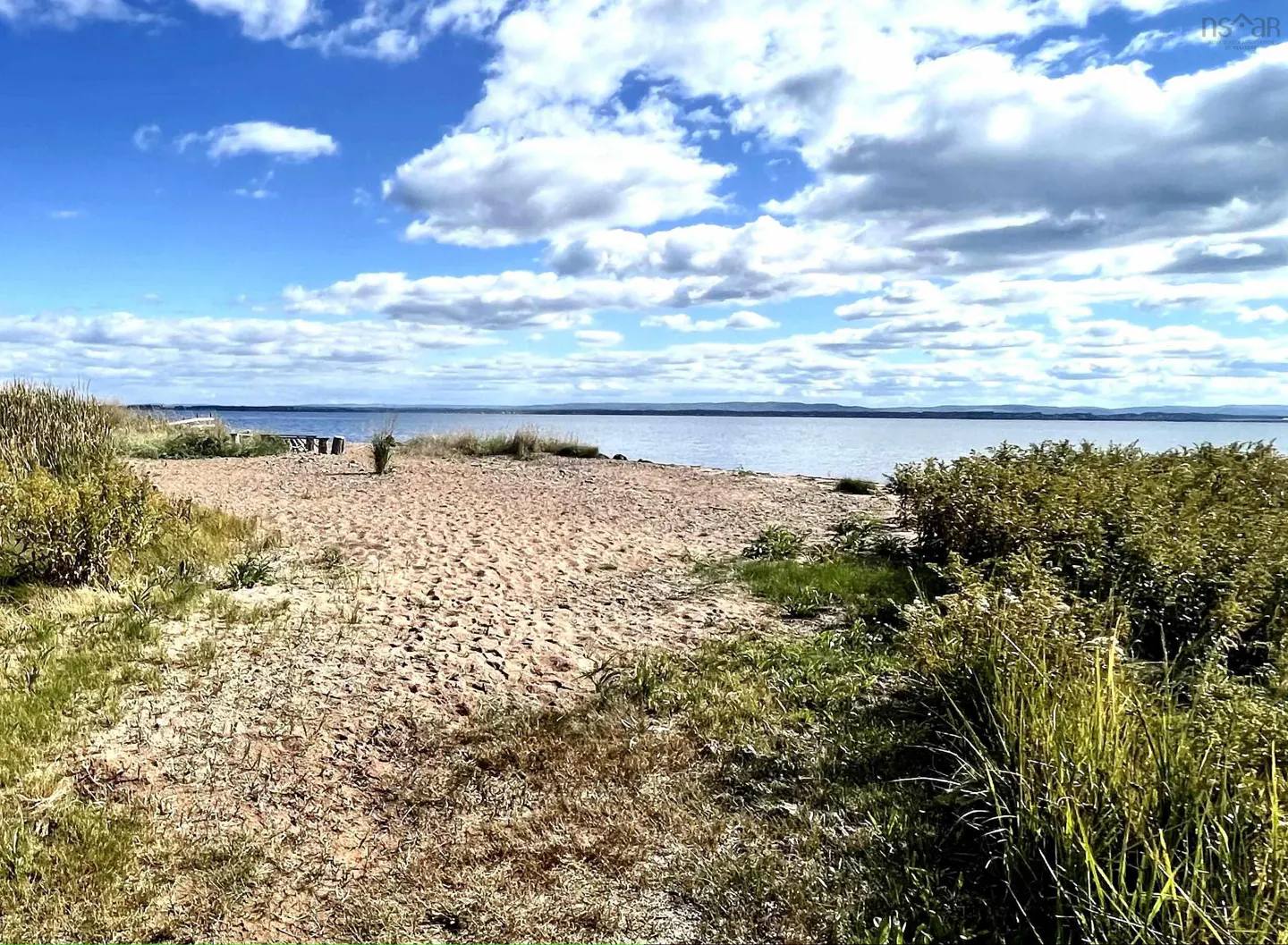 Sandy beach with green grass, blue water, and a cloudy blue sky.