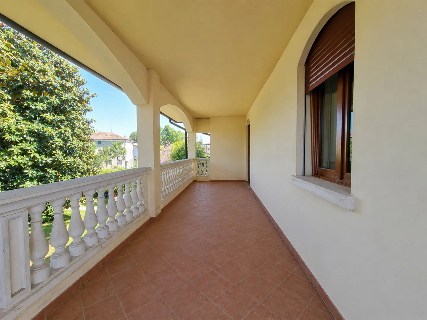 Long, covered balcony with a white balustrade, terracotta tile floor, and arched window with brown shutters. Trees and buildings are visible in the background.