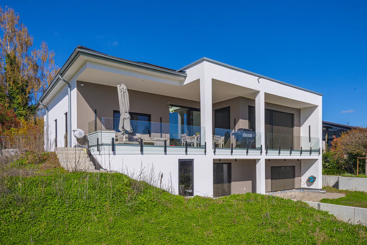 Two-story modern home with white and tan exterior, glass balcony, and green lawn under a clear blue sky.