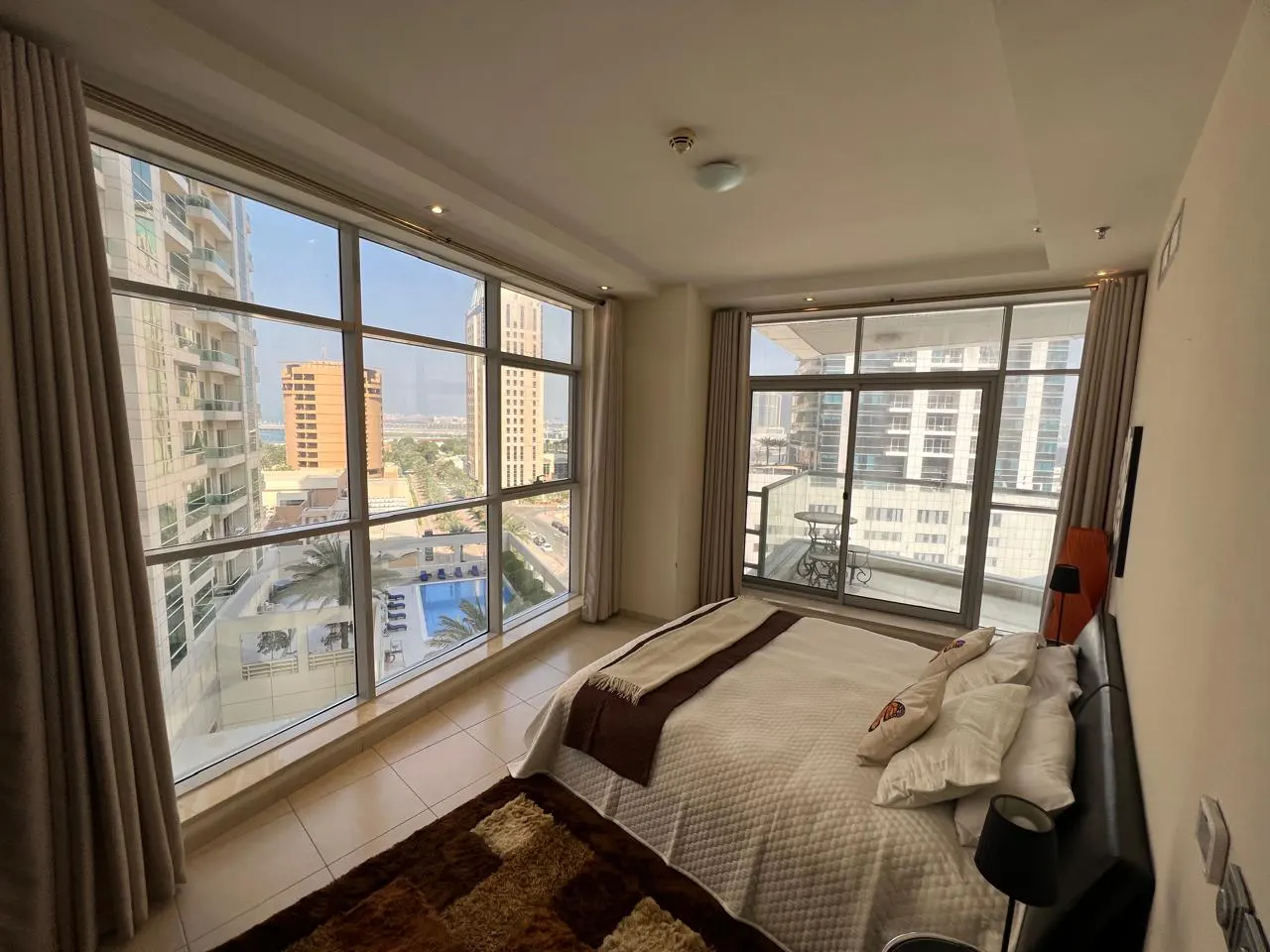 Bedroom with a large window view of city buildings, a pool, and a balcony. The bed has white linens and brown accents.