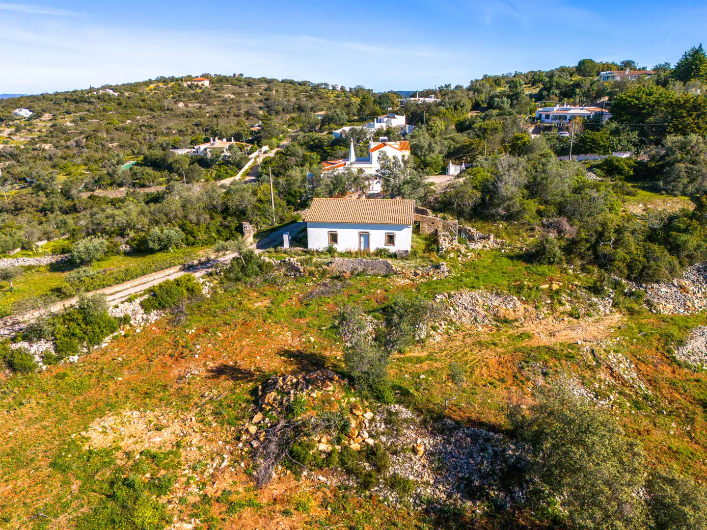 Aerial view of a small, white house with a terracotta roof, nestled in a green, hilly landscape under a blue sky.