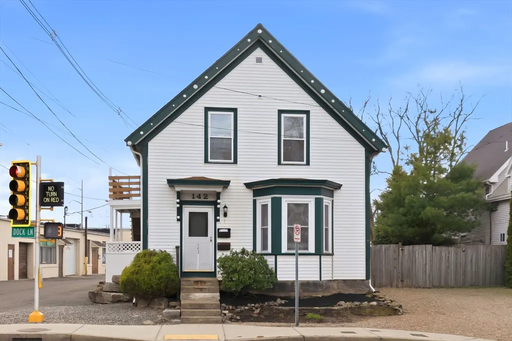 A two-story white house with green trim and the number 142 above the front door. A traffic light is on the left.