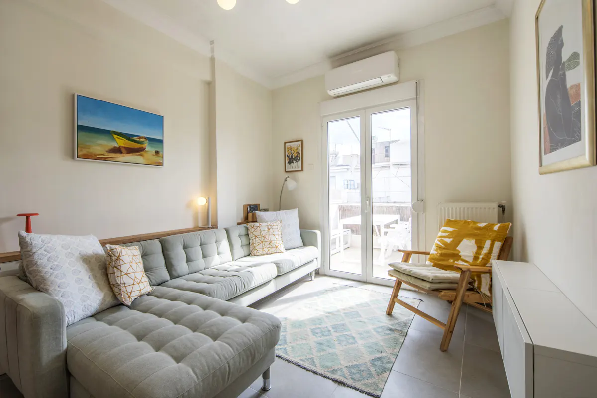 Bright living room with a light green sectional sofa, wooden chair with yellow pillow, and balcony access. Artwork adorns the walls.