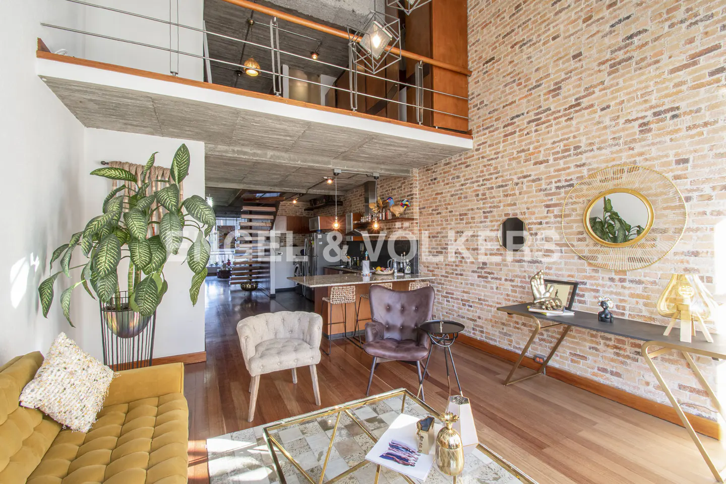 A modern living room with exposed brick, a yellow couch, and a glass coffee table. A loft overlooks the space.