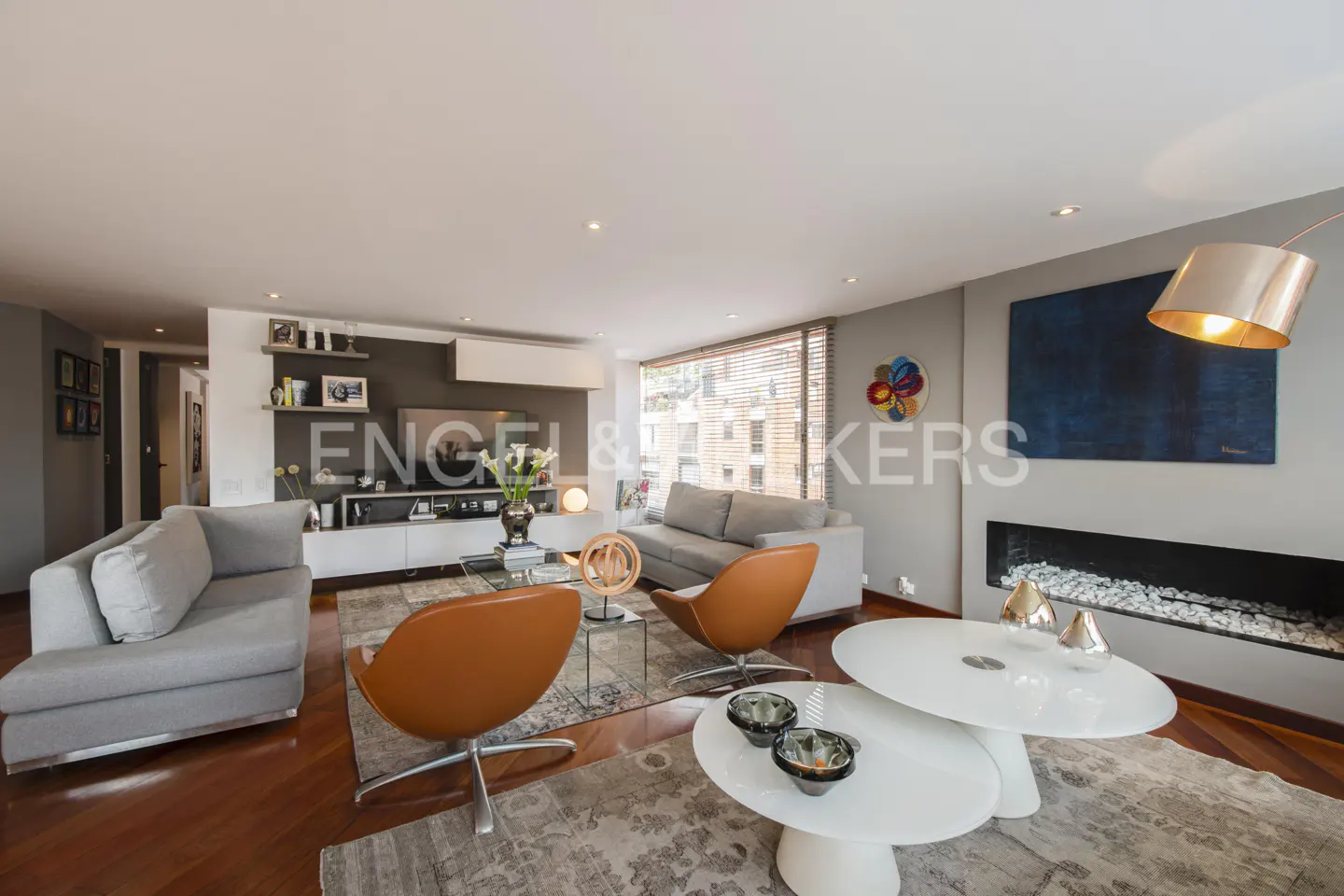 Bright living room with hardwood floors, gray sofas, and orange swivel chairs. A modern fireplace and blue artwork add style.
