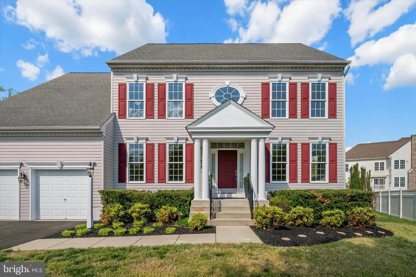 Beige two-story house with red shutters, white trim, and a red front door under a blue sky with white clouds.