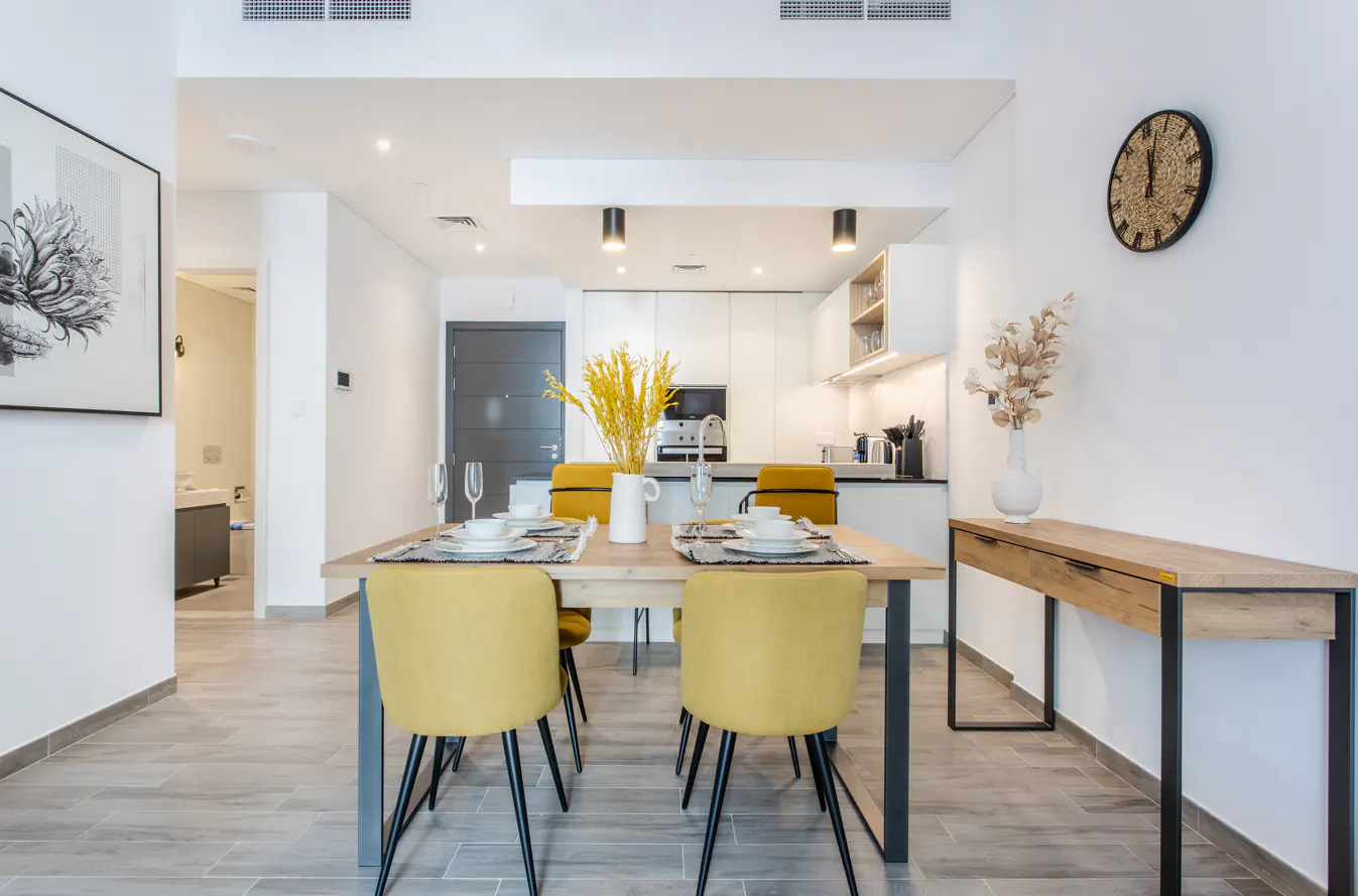 Bright dining area with a wooden table set for four, yellow chairs, and a white kitchen in the background. A console table sits against the wall.
