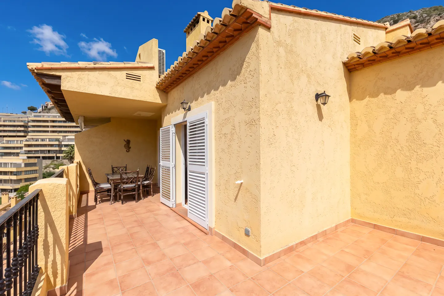 A sunny terrace with a table and chairs, terracotta tiles, and a yellow stucco wall with white shuttered doors.
