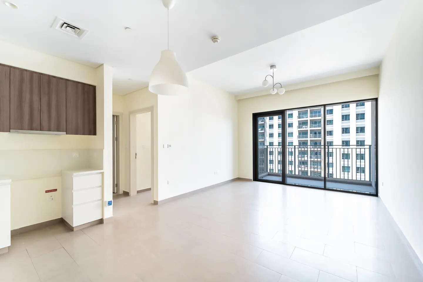 Bright, empty apartment with tile floors, white walls, and dark wood cabinets. A sliding glass door leads to a balcony with a city view.