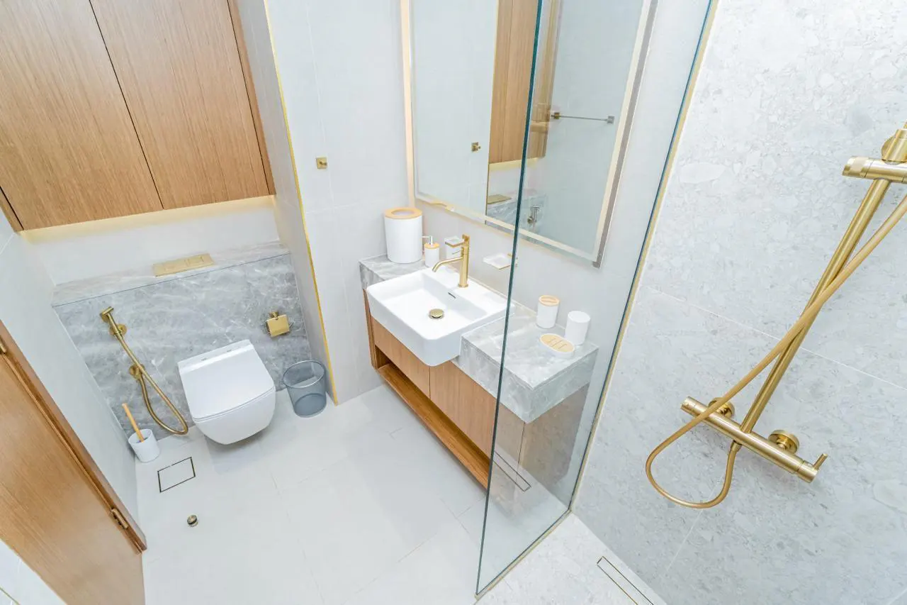 Bright bathroom with white tiles, a white toilet, and a sink with gold fixtures. A glass shower door is on the right.
