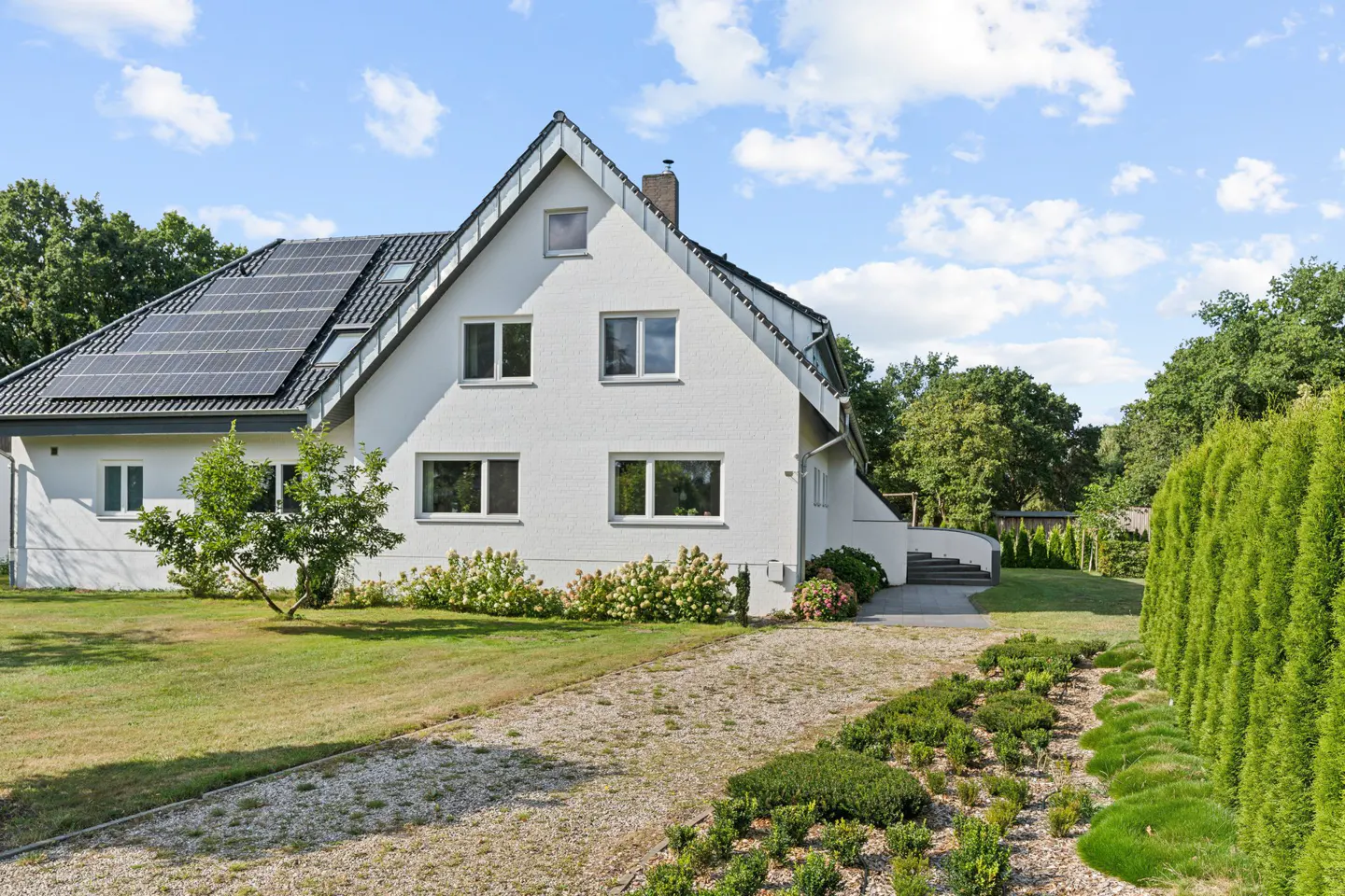 A white two-story house with solar panels on the roof, a green lawn, and a gravel driveway.