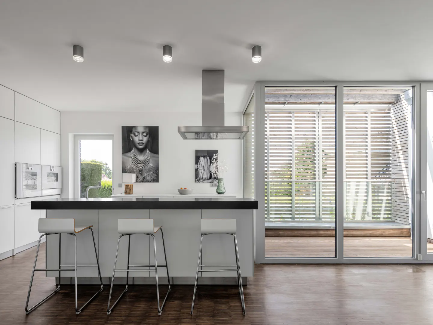 Bright, modern kitchen with white cabinets, black countertop, and three stools. Large windows with blinds overlook a green outdoor space.