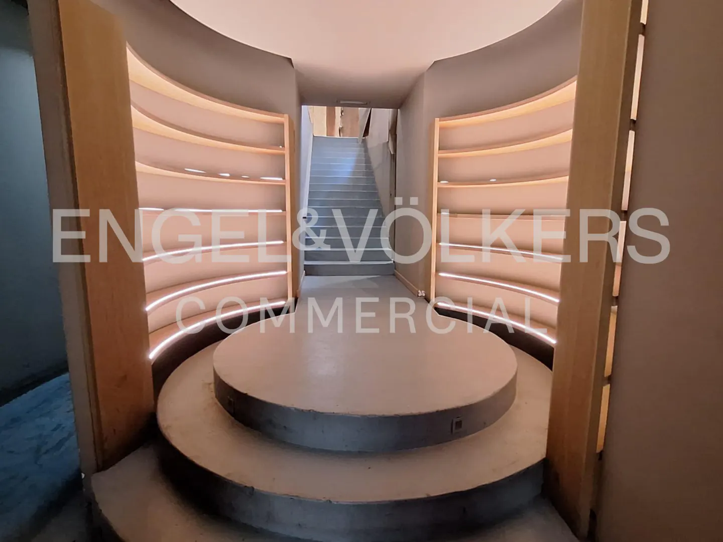 Interior view of a modern commercial space with a raised circular platform and illuminated curved shelves leading to stairs.