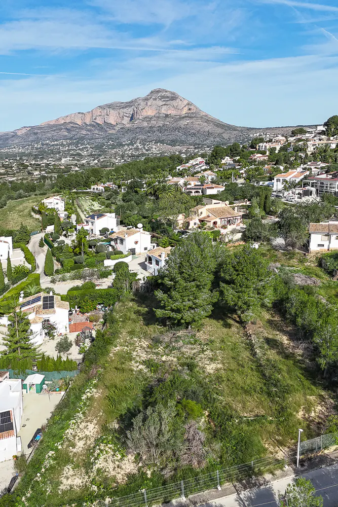 A scenic view of a grassy lot with trees, houses, and a mountain under a blue sky.