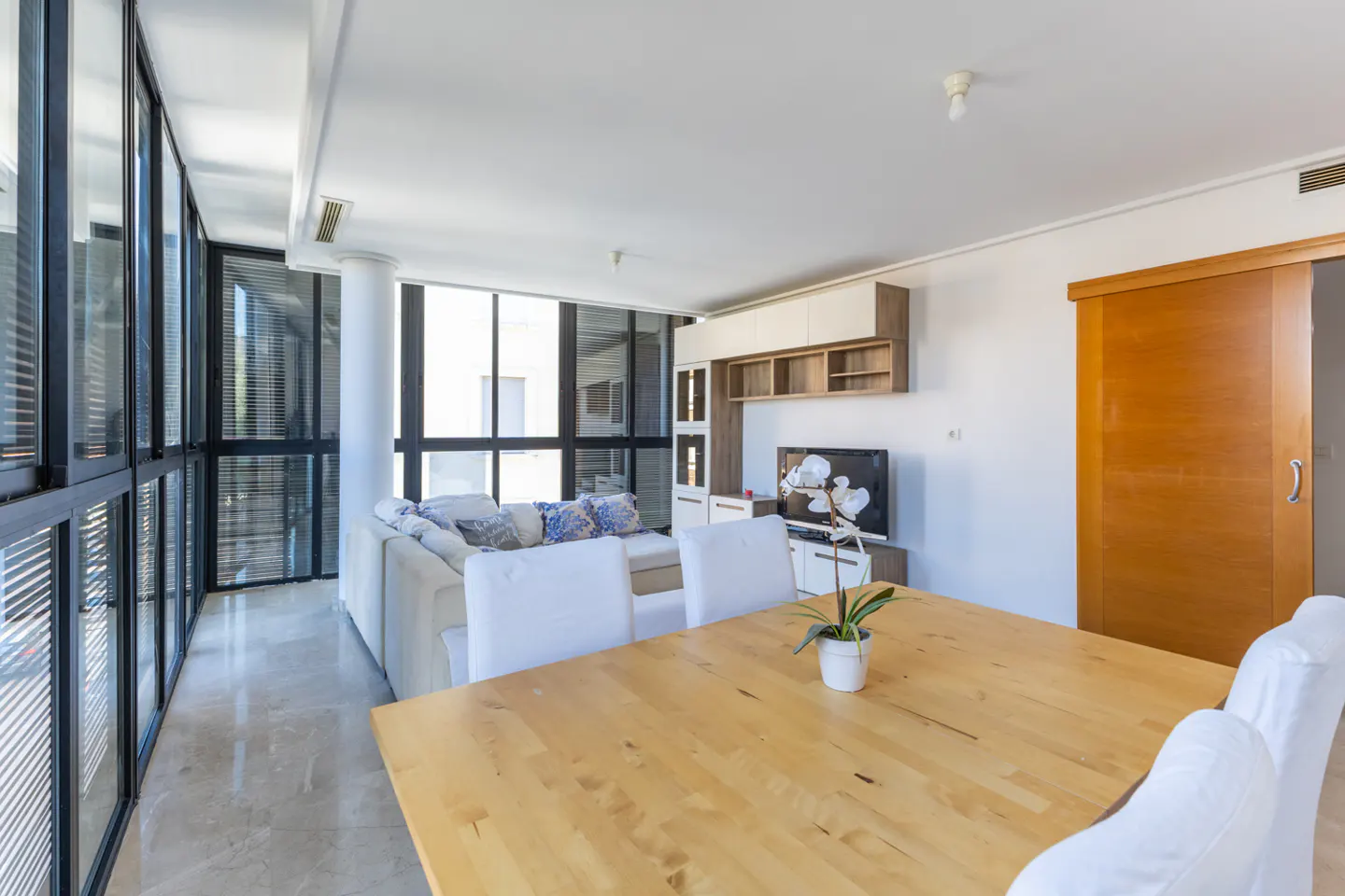 Bright living room with a wood table, white chairs, and a large window wall. A white sofa and TV are in the background.