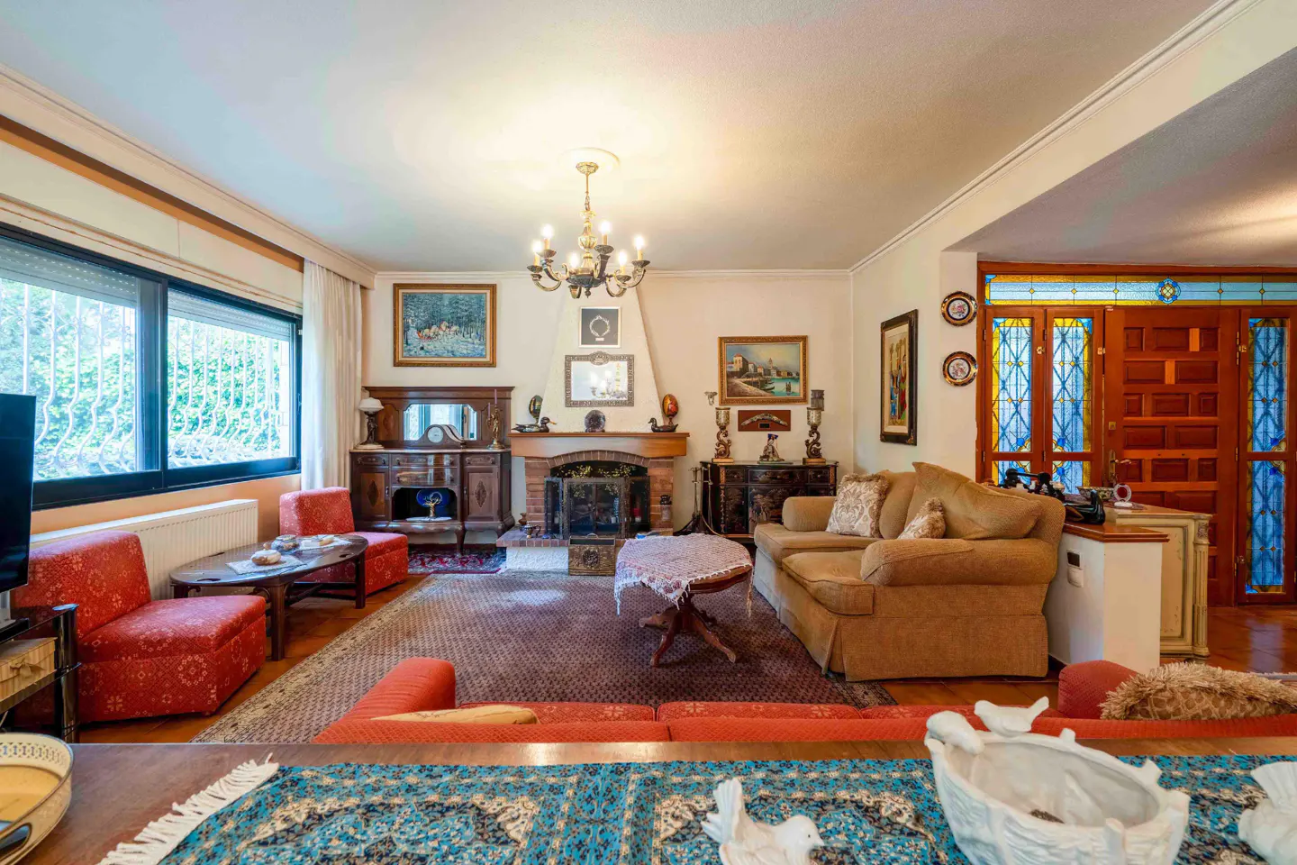 Cozy living room with a brick fireplace, tan sofa, red patterned chairs, a chandelier, and stained-glass wooden doors.