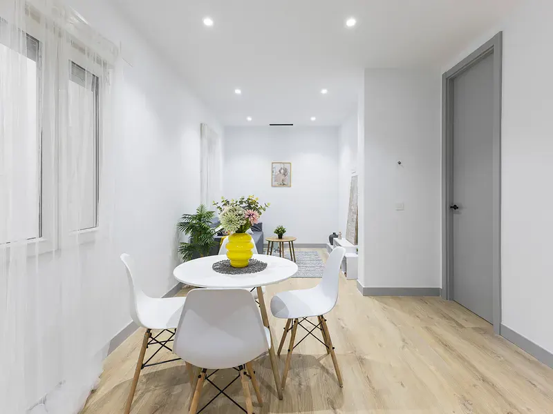 Bright, modern apartment interior with a round white table, four chairs, and a yellow vase of flowers. Wood floors and a gray door add contrast.
