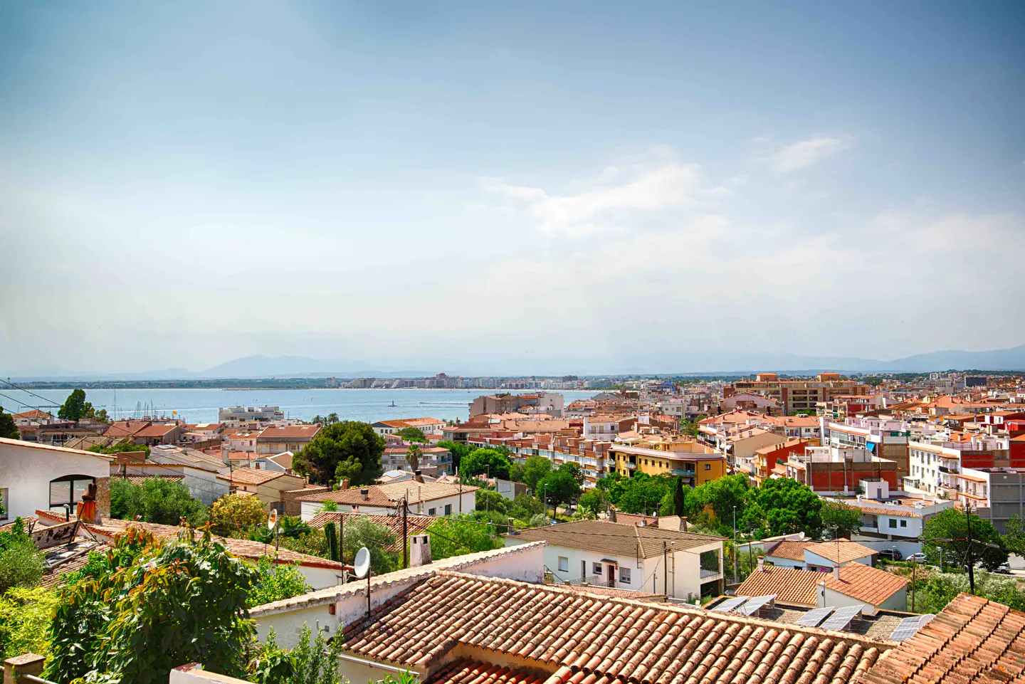 High-angle view of a coastal town with terracotta-roofed buildings, lush green trees, and a blue sea under a clear sky.