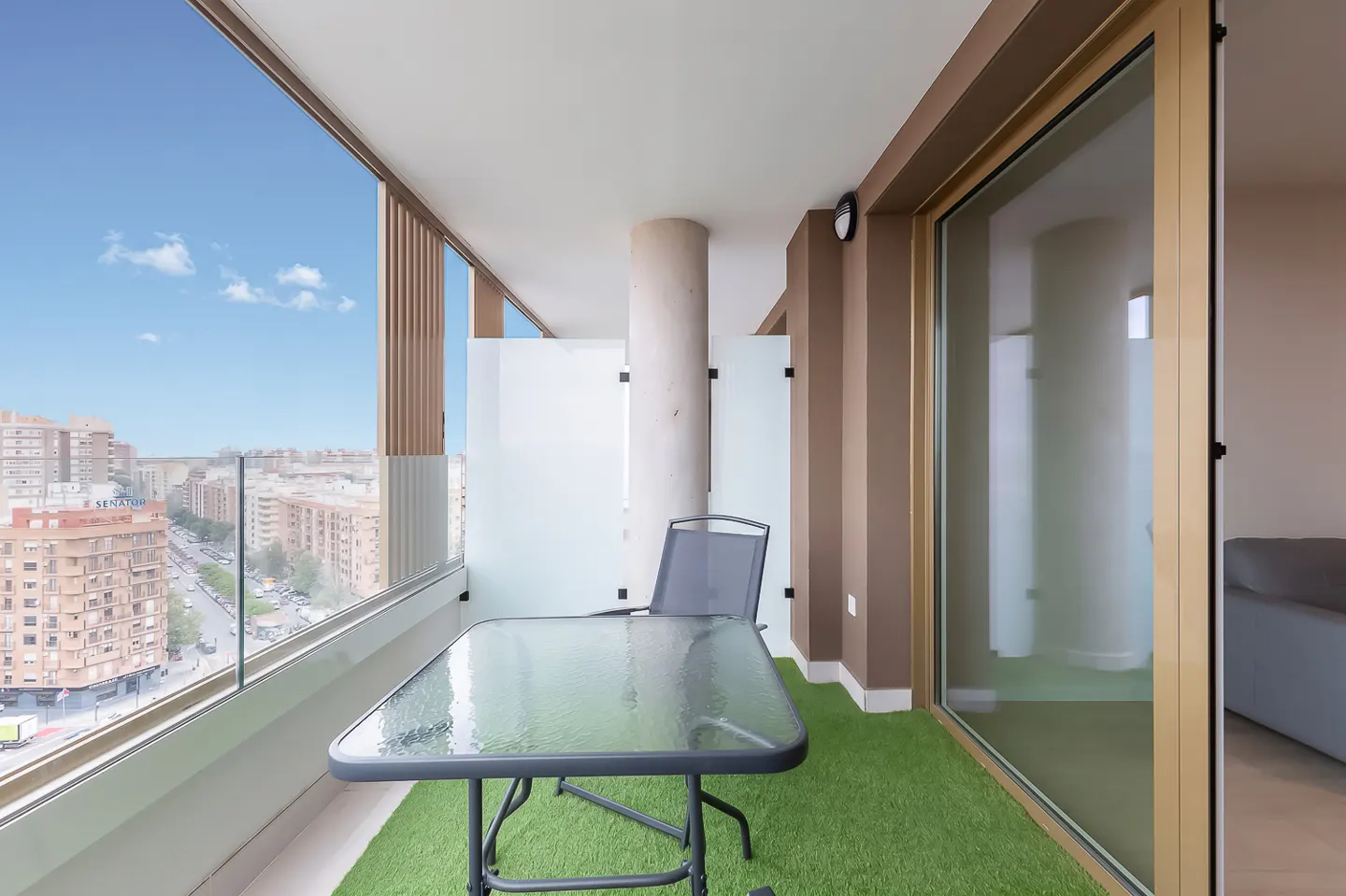 Balcony with glass table, chair, and artificial grass. City view with blue sky. Interior with sliding glass door.