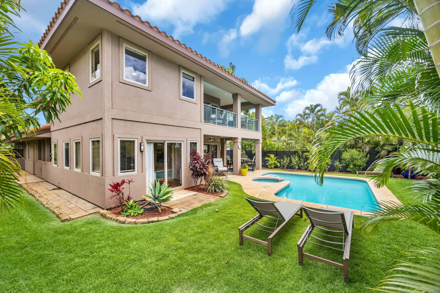 Two-story beige house with a backyard pool, lounge chairs on the lawn, and lush tropical landscaping under a blue sky.