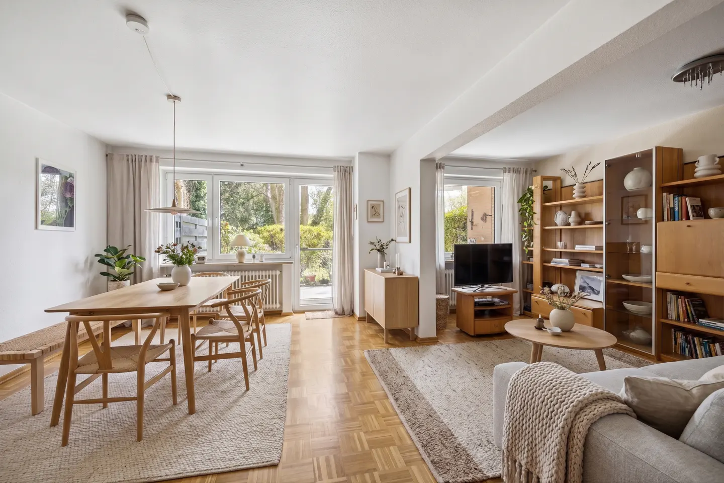 Bright, open-plan living and dining area with light wood furniture and parquet flooring. Large windows overlook a green garden.