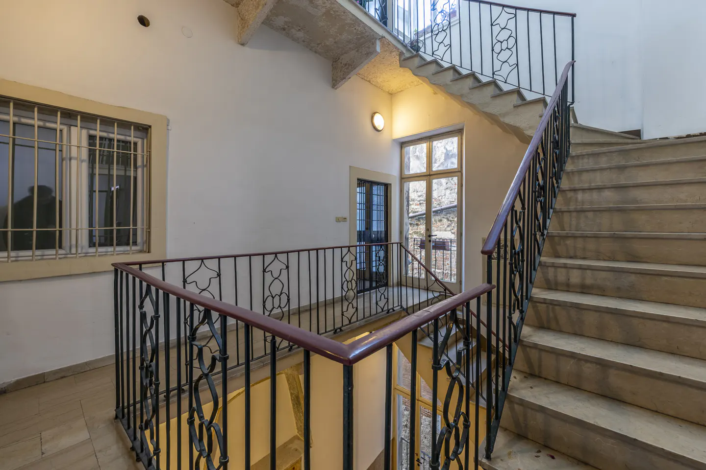 Interior view of a stairwell with stone steps, black iron railings, and a window with bars. The walls are white, and a light fixture illuminates the landing.
