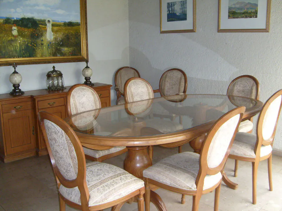 Dining room with a glass-topped table, six chairs, and a wooden sideboard with lamps and a painting.
