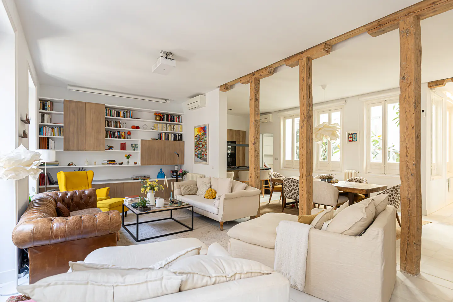 Bright, open-concept living and dining area with white walls, wooden beams, and neutral furniture. Bookshelves and a yellow chair add pops of color.
