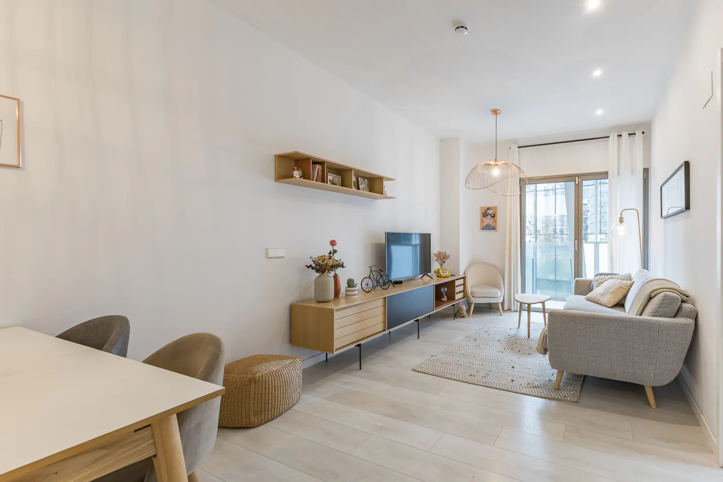 Modern living room with light wood floors, a grey sofa, a long media console, and a dining area with white table and chairs.