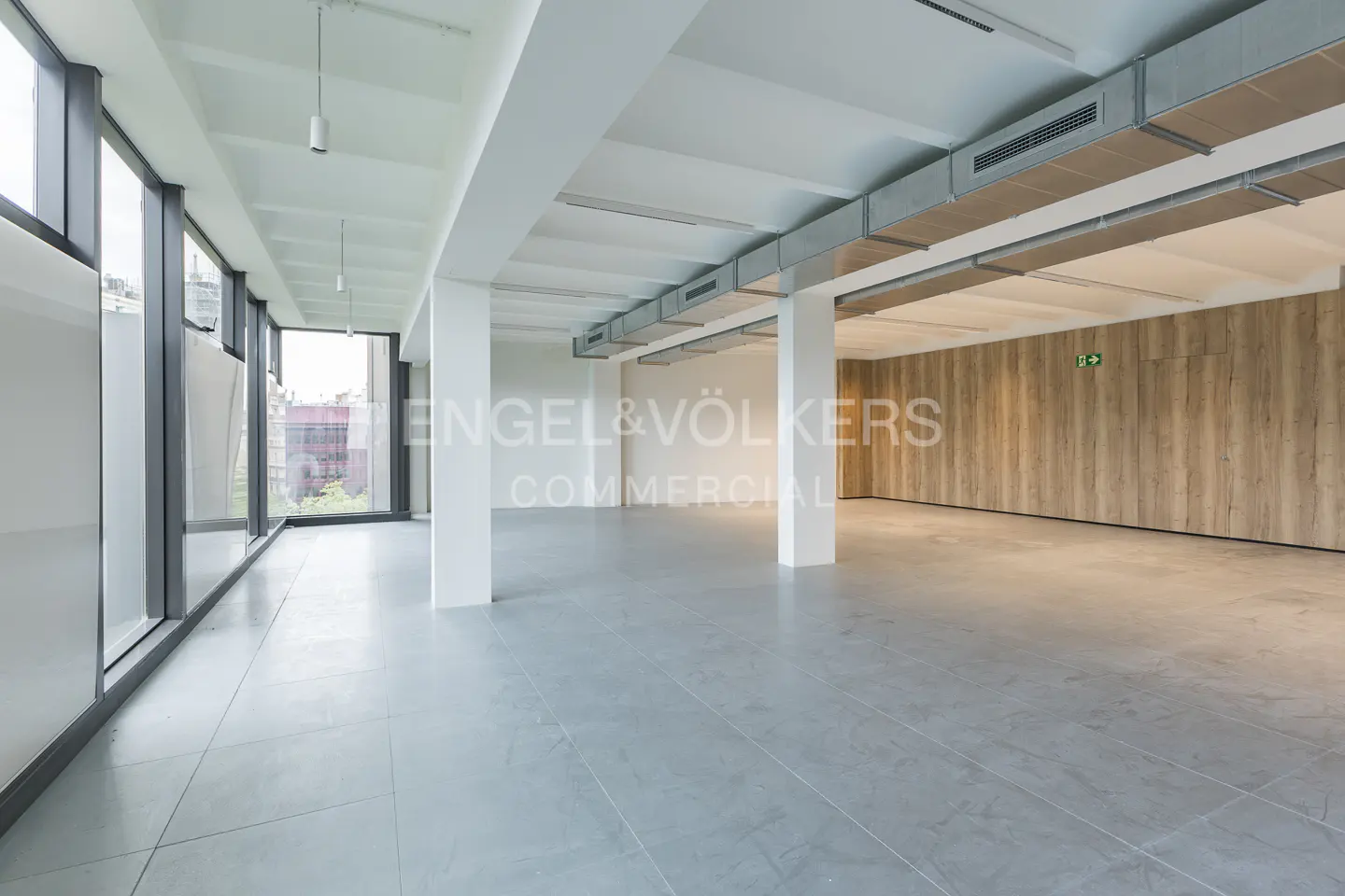 Empty commercial space with gray tile floor, white columns, and large windows. Wood-paneled wall with green exit sign in the background.