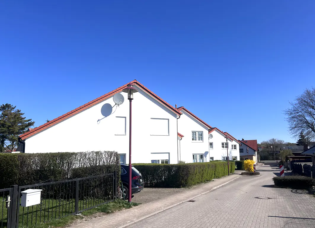 Row of white houses with red roofs under a clear blue sky. Hedges and a black fence line the street. A car is parked near a lamppost.