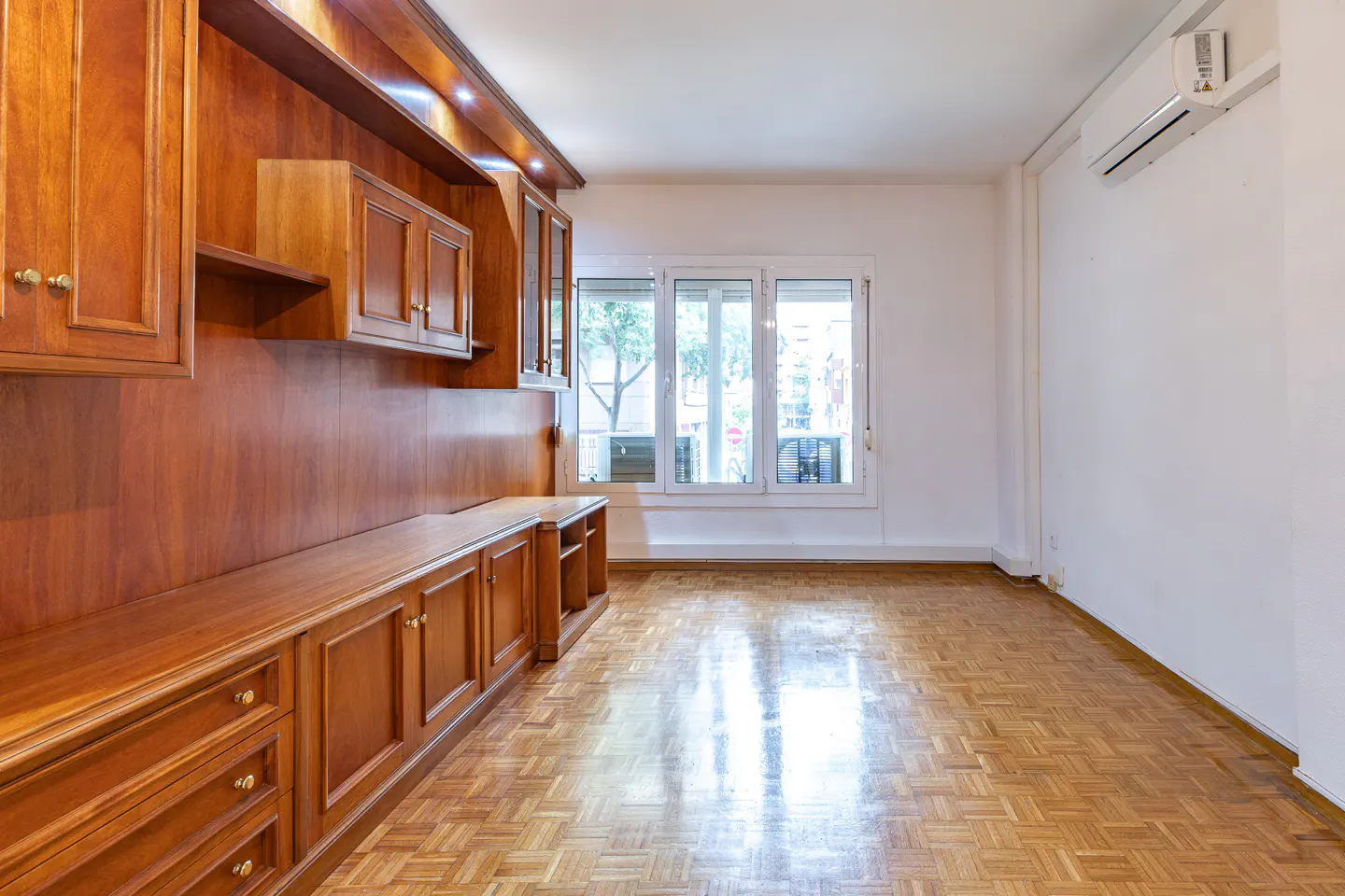 Empty room with wooden parquet flooring, built-in cherry wood cabinetry, white walls, and a window overlooking the street.