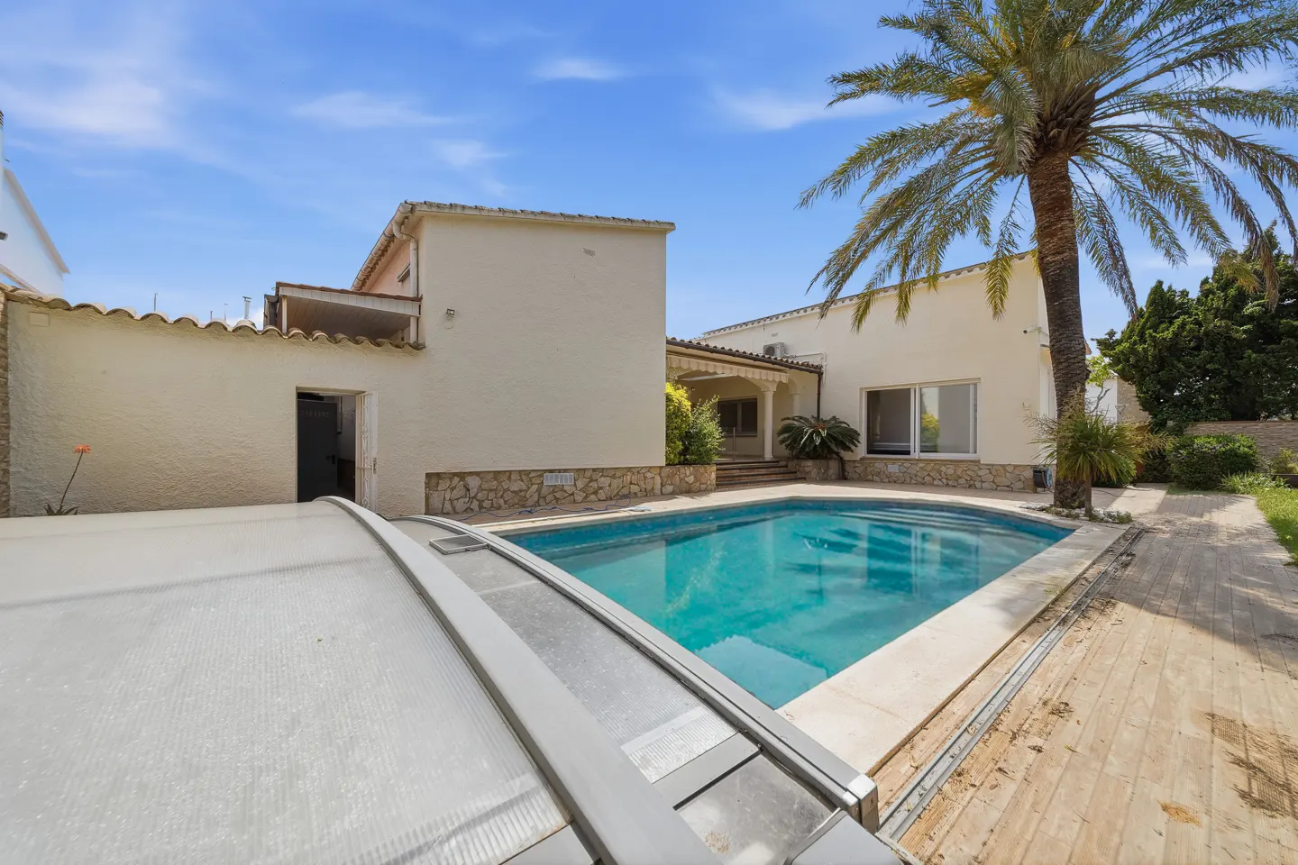 Backyard view of a cream-colored house with a turquoise pool, palm tree, and pool cover on a sunny day.