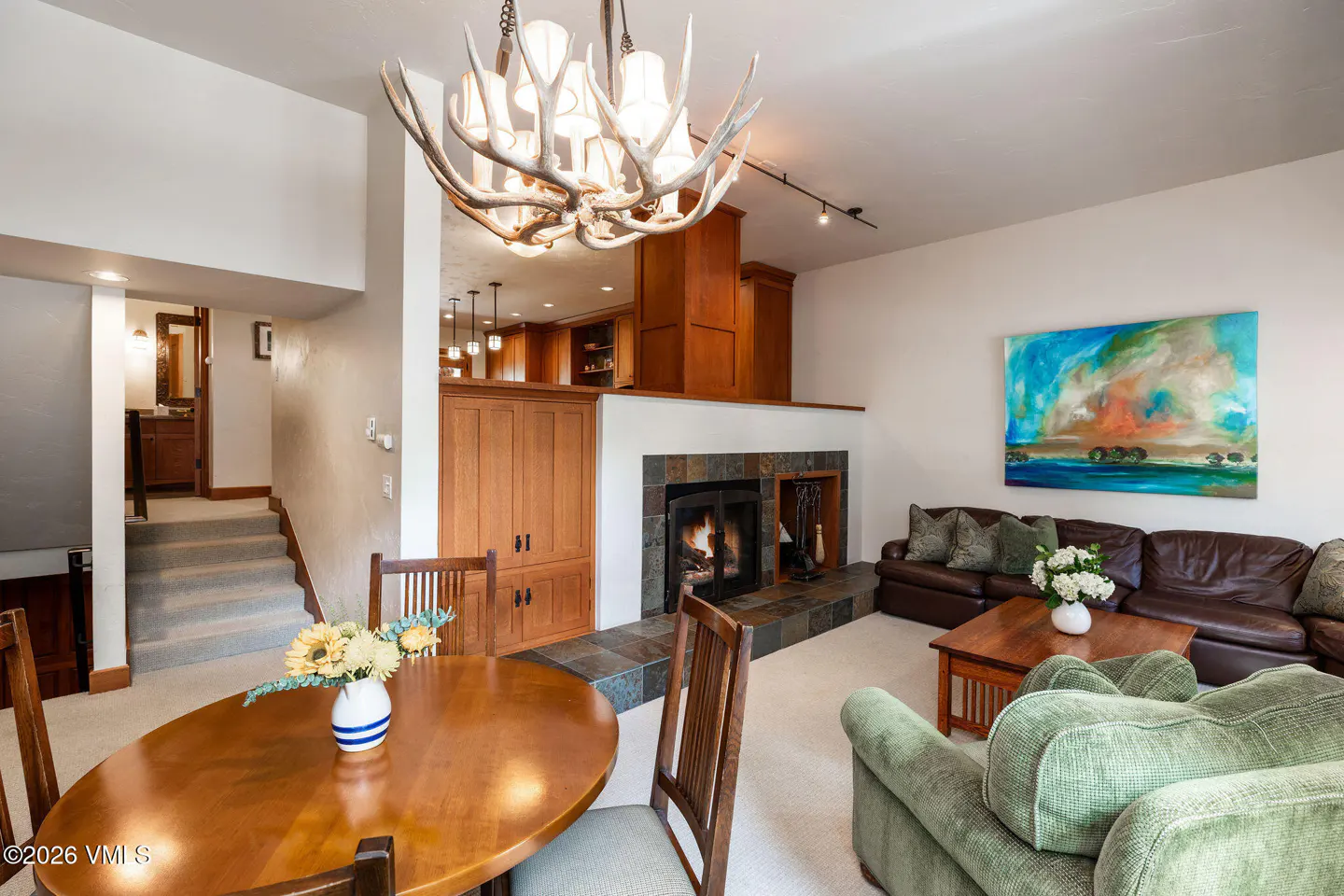 Living room with a fireplace, brown leather sofa, green armchair, and a wooden dining table with a flower vase. A chandelier made of antlers hangs from the ceiling.