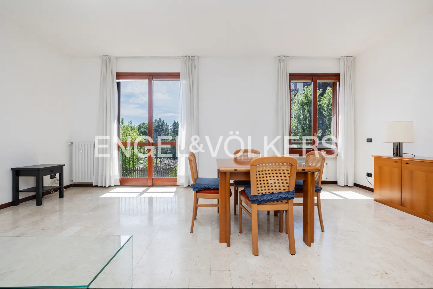 Bright dining room with marble floors, a wooden table and chairs, and large windows with white curtains. A wooden cabinet and black side table are also visible.