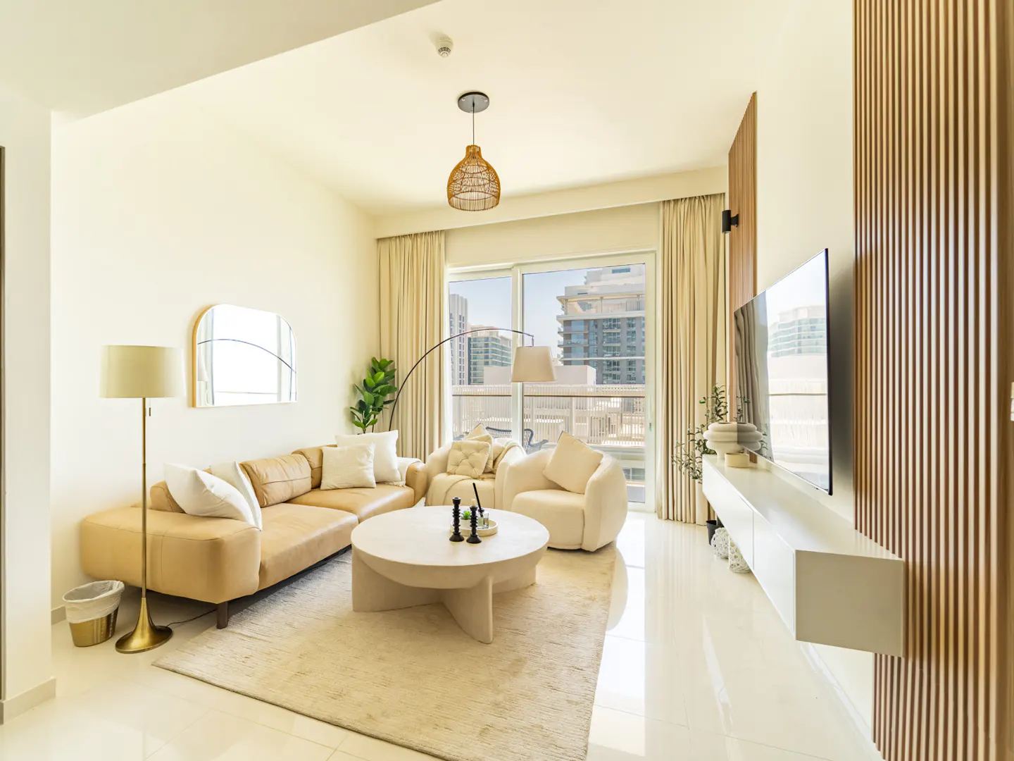 Modern living room with a beige sofa, round coffee table, white armchair, and wall-mounted TV, overlooking city buildings.